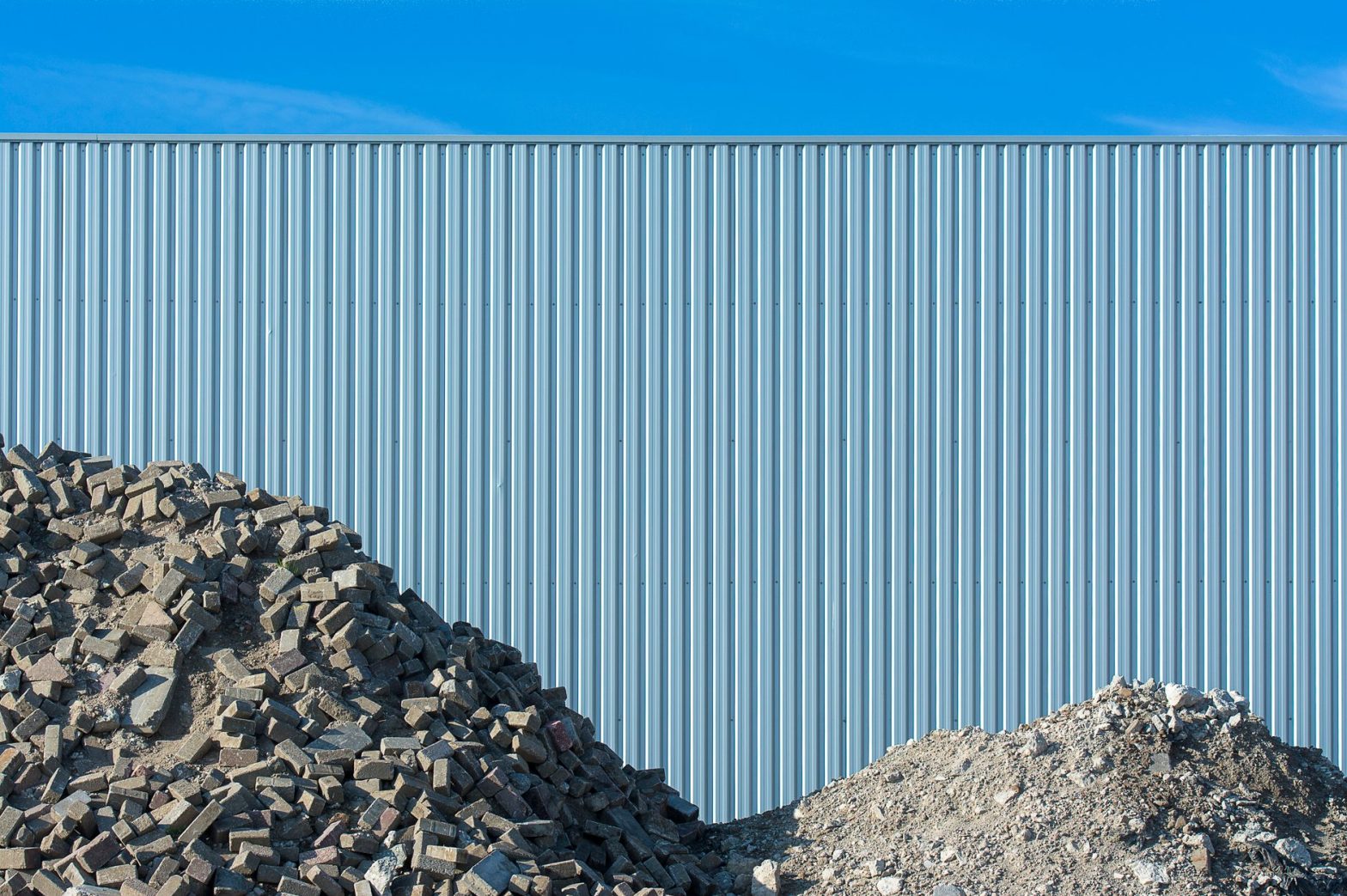 A pile of rubble in front of a large corrugated metal fence under a clear blue sky. — Asbestos Test for Property Owners, Tena