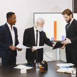 Three businessmen discussing documents in a conference setting with a presentation screen. — Find the Best Forex Options for