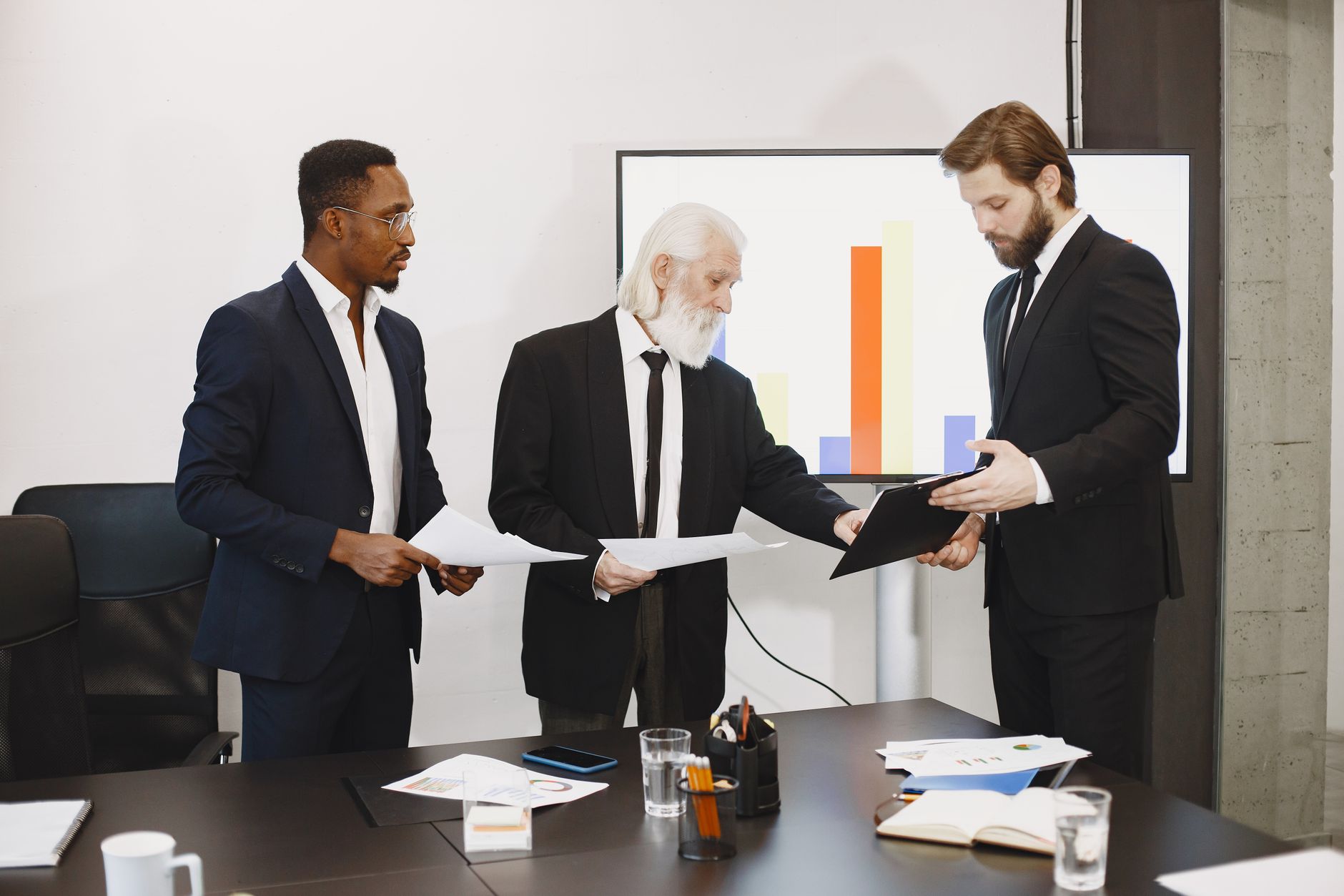 Three businessmen discussing documents in a conference setting with a presentation screen. — Find the Best Forex Options for