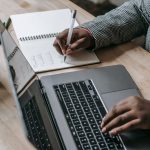 Crop unrecognizable African American female in formal wear writing notes in notepad while working on laptop — Praktyczne zast
